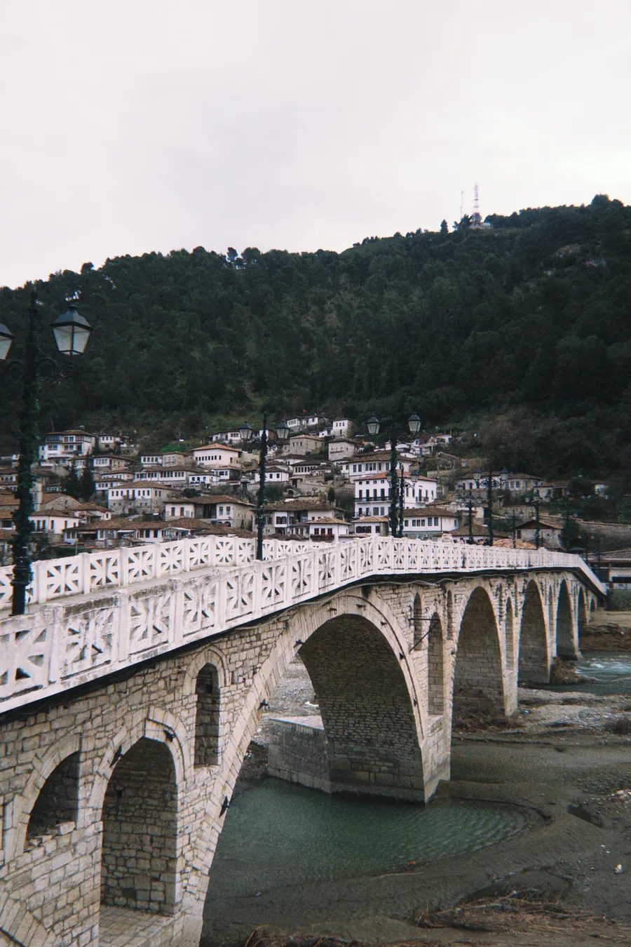 The old stone Gorica Bridge over the Osum River, with the white houses of Mangalem on the hillside behind