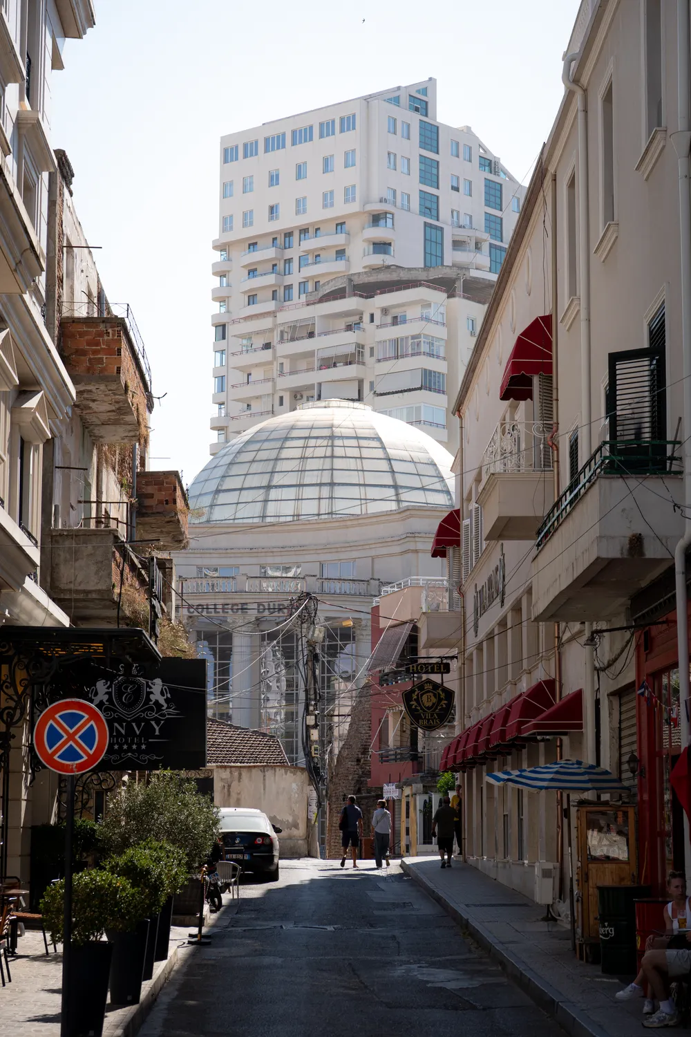 A narrow old-town street in Durrës, with a glass dome and modern tower blocks rising behind the older buildings