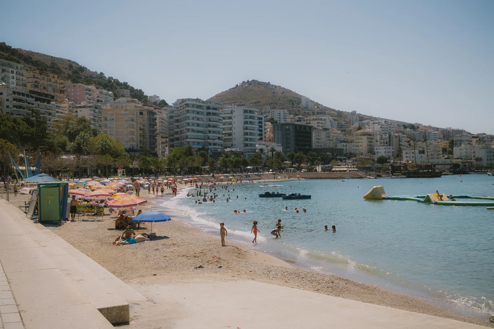 Sarandë’s town beach in summer: umbrellas and swimmers along the bay, with the apartment blocks and hills behind