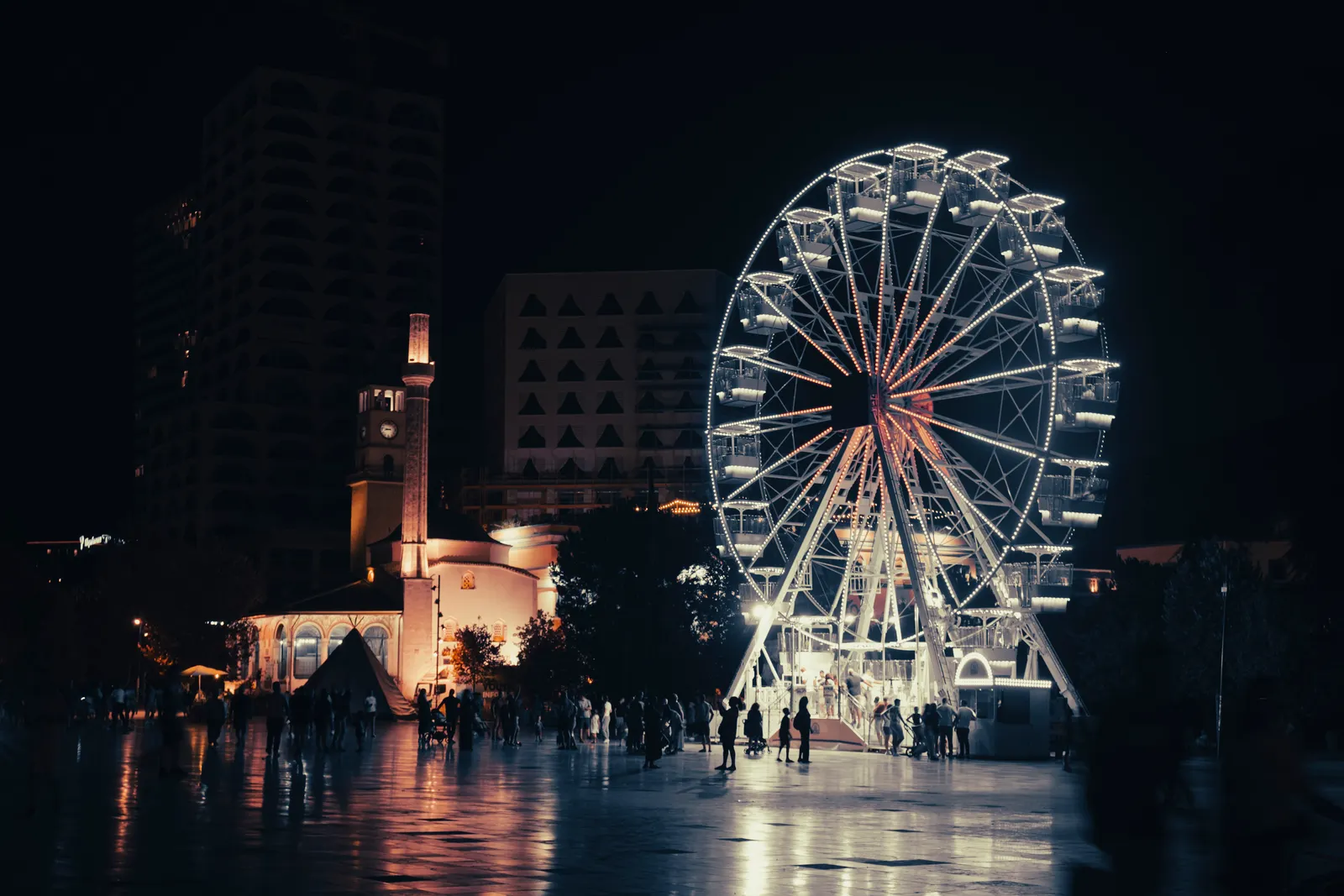Tirana at night: the Ferris wheel and Et’hem Bey Mosque lit up on Skanderbeg Square, people crossing the square